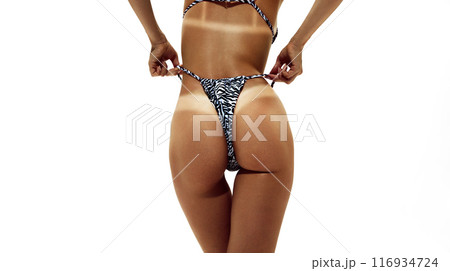 Woman pulls her animal-printed bikini, showing off distinct tan lines on her well-tanned skin against white studio background. 116934724