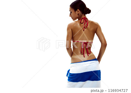 Woman holds towel against her buttocks, her glistening, deeply tanned skin showing clear tan lines against white studio background. 116934727
