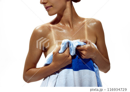 Woman wraps herself in towel, revealing stark tan lines on her deeply tanned skin against white studio background. 116934729