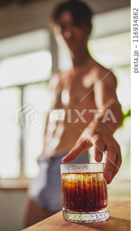 Shirtless man in denim shorts reaches for glass of iced dark beverage on table, with blurred background of sunlit room. 116934862