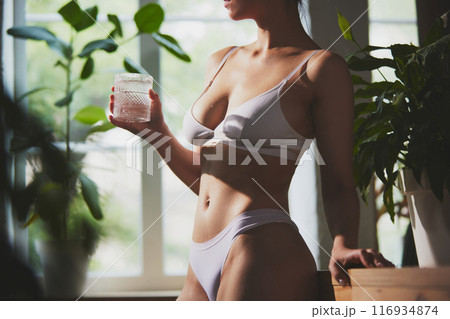 Fit woman dressed white inner wear stands by window, holding glass of water with ice, with soft natural light and greenery 116934874