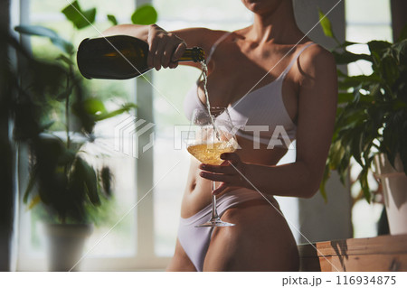 Woman in white lingerie pours champagne into glass standing in kitchen illuminated by natural light. Relaxed and elegant atmosphere. 116934875