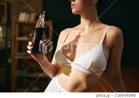 Cropped photo of young woman in white lingerie holding battle of soda serving with straw against blurred background. 116934899