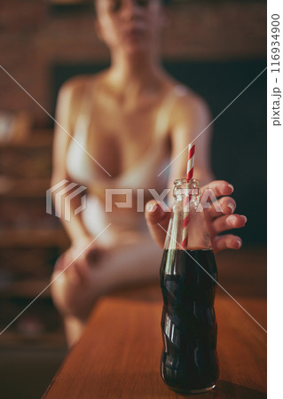 Young woman in white bikini sitting on wooden table and reaching to battle of soda serving with straw. Photo with selective focus. 116934900
