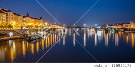 A view of the Rasin Embankment and Palacky Bridge in Prague, Czechia at dusk. The bridge is lit up, and the city lights reflect in the water. There are boats on the river, and the sky is a deep blue. 116935172