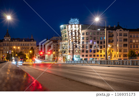 The Dancing House in Prague is illuminated by the city lights at dusk. The unique deconstructivist architecture stands out against the backdrop of traditional buildings. Light trails from passing cars 116935173