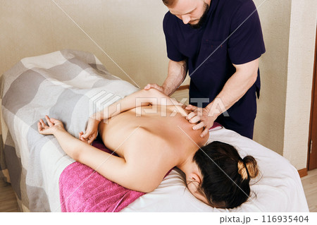 Masseur massages woman shoulder area while she lies face down on massage table. Masseur massages woman shoulder area while she lies face down on massage table. 116935404