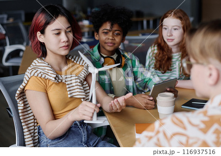 Asian girl holding wind turbine model discussing renewable energy sources with her classmates during Environmental Education lesson 116937516