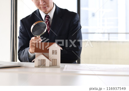 虫眼鏡で住宅模型を覗くスーツ姿の若い男性 116937549