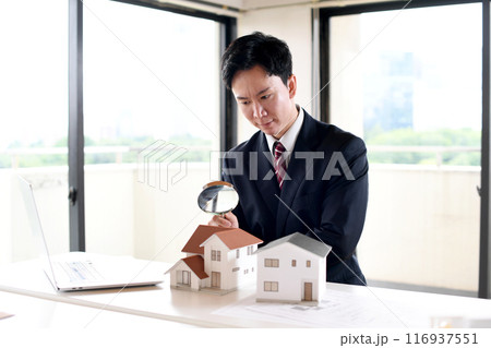 虫眼鏡で住宅模型を覗くスーツ姿の若い男性 116937551