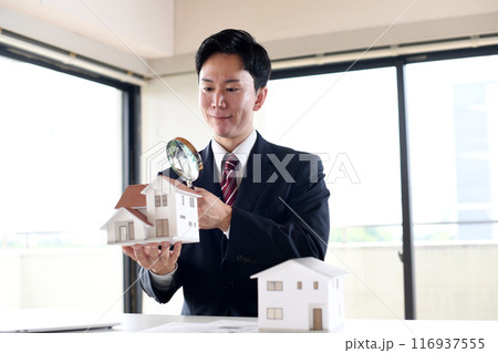 虫眼鏡で住宅模型を覗くスーツ姿の若い男性 116937555