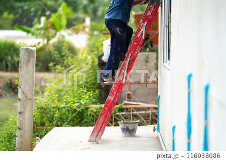 Man who wearing dark pants is climbing a red stairs to fix the house. Green shrub and broken pillar are on the background 116938086