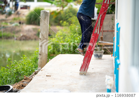 Man who wearing dark pants is climbing a red stairs to fix the house. Green shrub and broken pillar are on the background Man who wearing dark pants is climbing a red stairs to fix the house. Green shrub and broken pillar are on the background 116938087