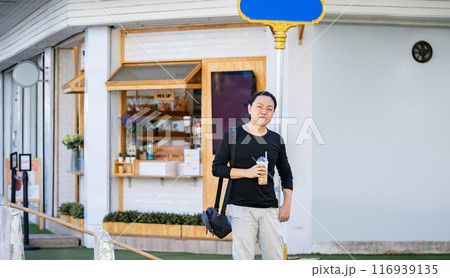 Asian man in the black t-shirt holds the bubble ball ice Milk Tea in front of the shop on the outdoor street footpath. Asian man in the black t-shirt holds the bubble ball ice Milk Tea in front of the shop on the outdoor street footpath. 116939135