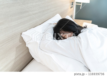 Asian Lady in white long arm t-shirt pajamas lie prone, lie on the stomach in bed with tried and exhausted mood and tone. 116939736