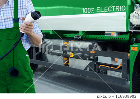 Driver in uniform holds electric vehicle charging plug. Electric truck in the background 116939902