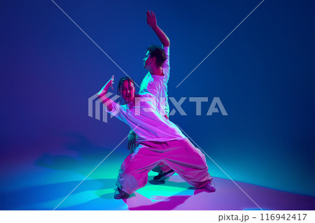 Cheerful, talented, positive young man and woman dancing hip-hop, making dynamic performance against purple studio background in neon light Cheerful, talented, positive young man and woman dancing hip-hop, making dynamic performance against purple studio background in neon light 116942417