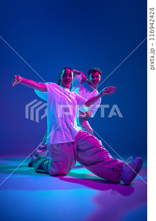 Dynamic image of artistic and energetic young man and woman, dancers performing hip-hop against purple studio background in neon light Dynamic image of artistic and energetic young man and woman, dancers performing hip-hop against purple studio background in neon light 116942428