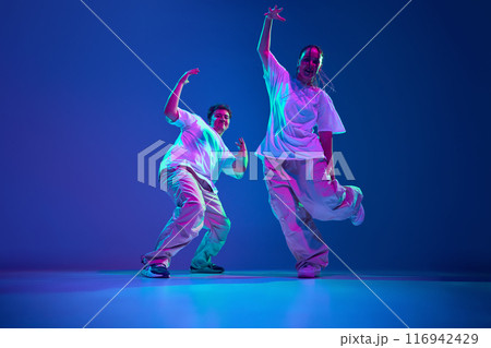 Dynamic image of artistic and energetic young man and woman, dancers performing hip-hop against purple studio background in neon light 116942429