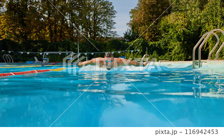Wearing goggles, swimmer powers through water with butterfly stroke, demonstrating strength and technique under bright sun. 116942453
