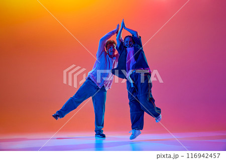 Young people, man and woman, dancers performing modern dance styles, dancing hip-hop against gradient studio background in neon light Young people, man and woman, dancers performing modern dance styles, dancing hip-hop against gradient studio background in neon light 116942457
