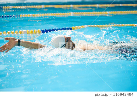 Fit man, swimmer, donning white cap, pushes forward in pool, his arm reaching out in strong freestyle stroke in sunlit pool outdoor. Fit man, swimmer, donning white cap, pushes forward in pool, his arm reaching out in strong freestyle stroke in sunlit pool outdoor. 116942494