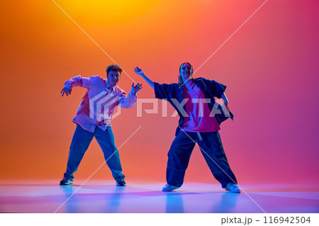 Dynamic image of young man and woman in casual oversized clothes dancing hip-hop against gradient studio background in neon light Dynamic image of young man and woman in casual oversized clothes dancing hip-hop against gradient studio background in neon light 116942504