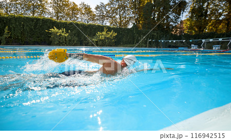 Athletic man wearing white swim cap, and goggles swims freestyle, utilizing yellow paddle to enhance his performance in sunny pool. Athletic man wearing white swim cap, and goggles swims freestyle, utilizing yellow paddle to enhance his performance in sunny pool. 116942515