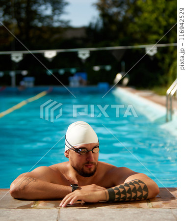Swimmer in white swim cap rests at pool's edge, wearing goggles and looking thoughtful against sunny outdoor backdrop. 116942529