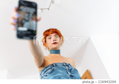 portrait of red-haired young woman wearing denim dress using phone 116943204
