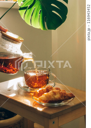 A hand pours tea from a glass teapot into a cup with croissants on a wooden table, a large green leaf in the background, vertical frame A hand pours tea from a glass teapot into a cup with croissants on a wooden table, a large green leaf in the background, vertical frame 116943452