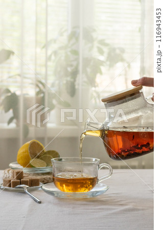 A hand pours brewed tea from glass teapot into a cup, with lemon slices and sugar cubes on the table near the window, cozy home environment A hand pours brewed tea from glass teapot into a cup, with lemon slices and sugar cubes on the table near the window, cozy home environment 116943453