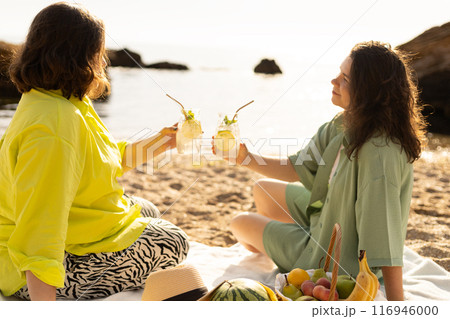 two girlfriends on a picnic with fruits and cocktails on the seashore 116946000