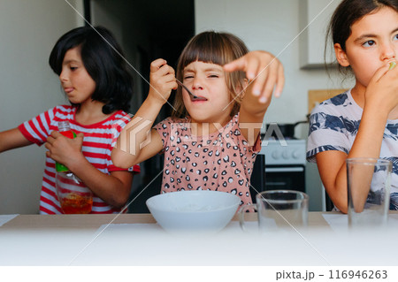 small angry child eating lunch at home at white table with two other children sitting on the side 116946263