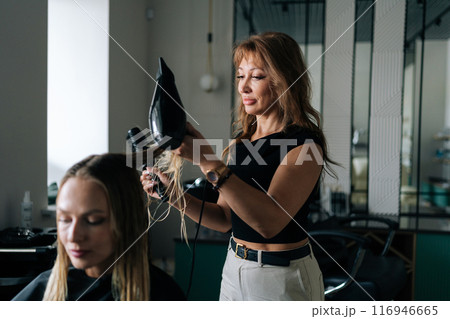 Medium shot of female hairstylist drying and straightening long blonde customer hair with comb and hairdryer in beauty salon. Pretty young woman lady client getting stylish hairdo done at salon. 116946665