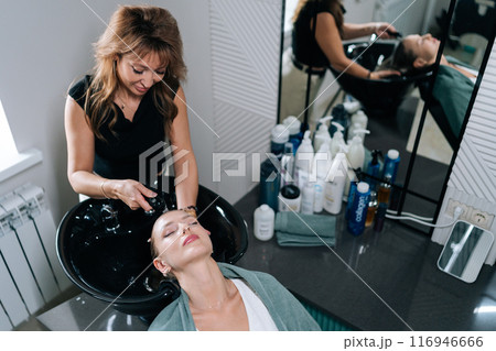 Top view of female hairstylist washing young woman client hair with warm water over sink in barbershop. Blonde female visiting beauty salon for modern haircut. Concept of fashion and beauty. Top view of female hairstylist washing young woman client hair with warm water over sink in barbershop. Blonde female visiting beauty salon for modern haircut. Concept of fashion and beauty. 116946666