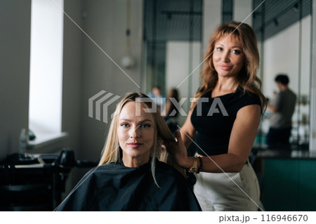 Portrait of professional hairdresser working on fashionable image for female customer, combing hair after applying paint with brush. Blonde young woman client getting hair dressed in beauty salon. 116946670
