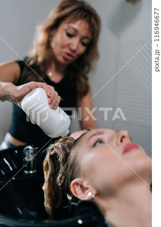 Vertical shot of professional hairstylist foaming shampoo on long blonde hair of beautiful female client. Skilled hairdresser washing head of young woman in beauty hairdressing salon. Vertical shot of professional hairstylist foaming shampoo on long blonde hair of beautiful female client. Skilled hairdresser washing head of young woman in beauty hairdressing salon. 116946677