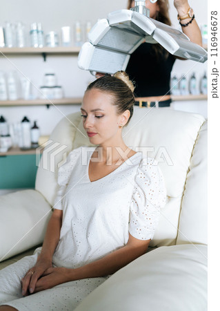 Vertical portrait of woman client with blonde hair having LED light hair PDT treatment at luxury aesthetic clinic. Female specialist adjusting innovation device for photodynamic therapy procedure Vertical portrait of woman client with blonde hair having LED light hair PDT treatment at luxury aesthetic clinic. Female specialist adjusting innovation device for photodynamic therapy procedure 116946678