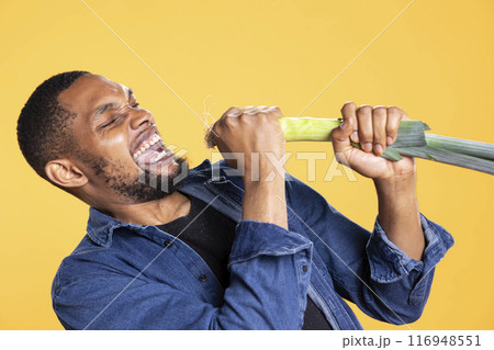 African american talented relaxed guy mimic singing on a leek pretending it is a microphone, advertising vegan nutrition and having fun. Confident silly person being funny fooling around. African american talented relaxed guy mimic singing on a leek pretending it is a microphone, advertising vegan nutrition and having fun. Confident silly person being funny fooling around. 116948551