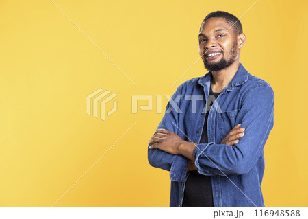 Portrait of confident african american person posing in studio, standing with arms crossed against yellow background. Enthusiastic relaxed guy smiling in front of the camera, masculine figure. Portrait of confident african american person posing in studio, standing with arms crossed against yellow background. Enthusiastic relaxed guy smiling in front of the camera, masculine figure. 116948588