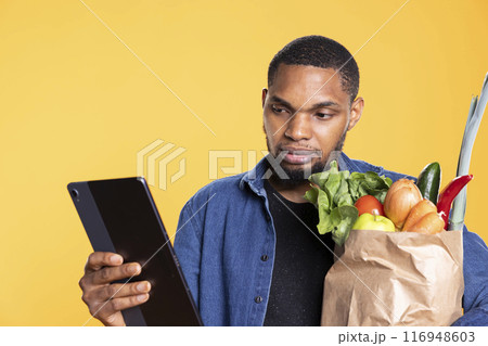 African american person checks his social media apps in studio, carrying a paper bag full of groceries from the grocery store. Happy relaxed man having fun browsing the internet, eco friendly. African american person checks his social media apps in studio, carrying a paper bag full of groceries from the grocery store. Happy relaxed man having fun browsing the internet, eco friendly. 116948603