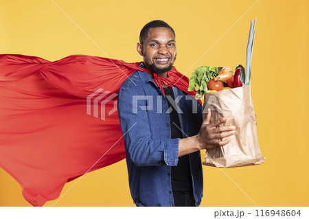 Superhero model with red cape posing with a bag of bio groceries, promoting sustainable lifestyle and locally grown veggies. Young person showcasing zero waste concept in studio. Superhero model with red cape posing with a bag of bio groceries, promoting sustainable lifestyle and locally grown veggies. Young person showcasing zero waste concept in studio. 116948604