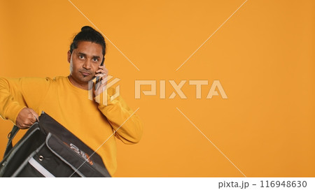 Man packing food in thermal backpack, fulfilling clients orders and answering phone ringing, studio backdrop. Indian person doing delivery to customers, discussing with them on cellphone, camera A 116948630