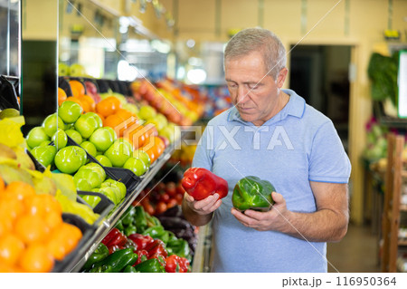 Focused elderly male pensioner in casual wear choosing local bell peppers during shopping in store 116950364