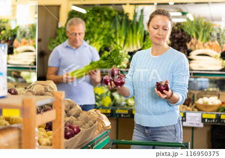 Busy casual adult woman pushing shopping cart and picking red onions in grocery shop 116950375