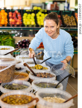 Portrait of focused adult European woman in casual clothes buying pickled olives in hypermarket Portrait of focused adult European woman in casual clothes buying pickled olives in hypermarket 116950507