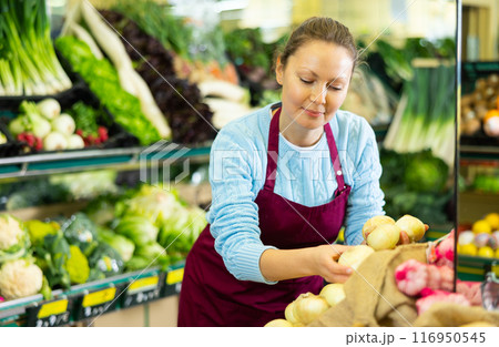 Middle-aged saleswoman putting onions on food stall in greengrocery 116950545