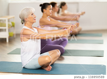 Women of different ages sit on mats and do pilates exercises with an expander in an gym 116950589