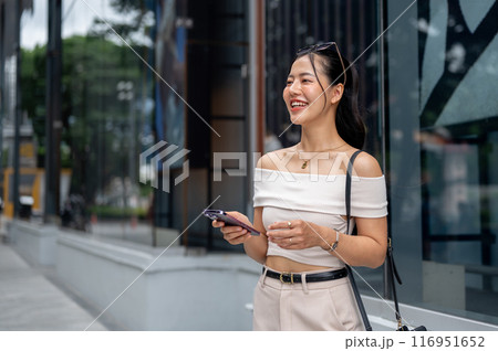 A beautiful Asian woman is standing in front of the shopping mall with a smartphone in her hand. 116951652